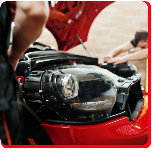 Technicians performing car maintenance on the engine and headlights of a red vehicle.