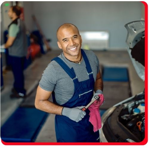 Smiling auto technician holding a wrench and cloth beside a car receiving an oil change.