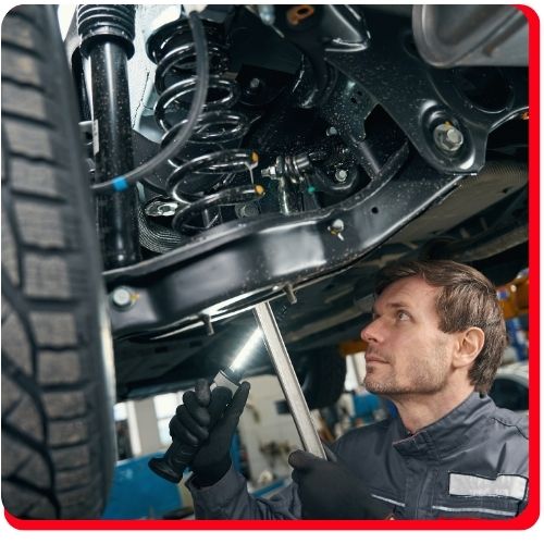 Mechanic inspecting a vehicle's suspension system with a flashlight and tool.